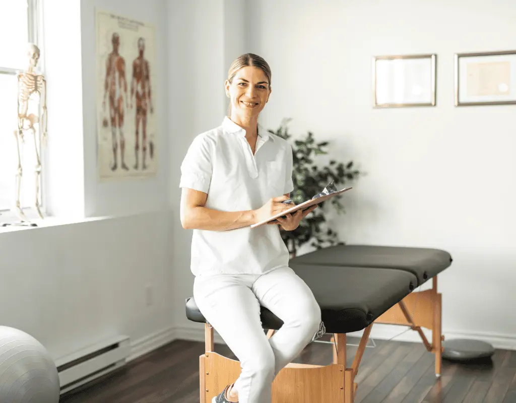 Female clinician in white seated at the edge of a treatment table holding a clipboard in a bright room with anatomy charts