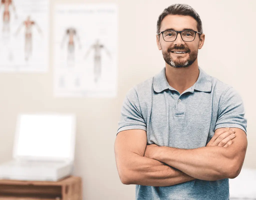 Bearded man in a blue polo shirt standing with arms folded in a treatment room with anatomy posters on the wall