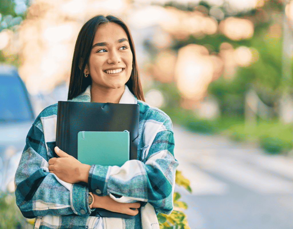 Smiling student standing outdoors holding a folder and tablet against a sunlit background