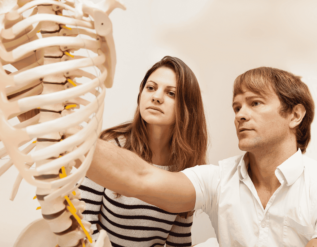 Woman demonstrating shoulder movement beside an anatomical skeleton model while a man observes