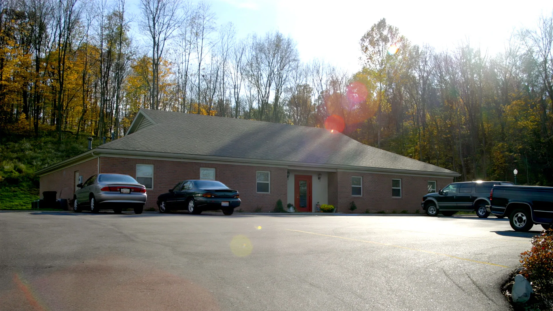 Brick single-story clinic behind a parking area, framed by autumn trees with sunlight flaring through the branches