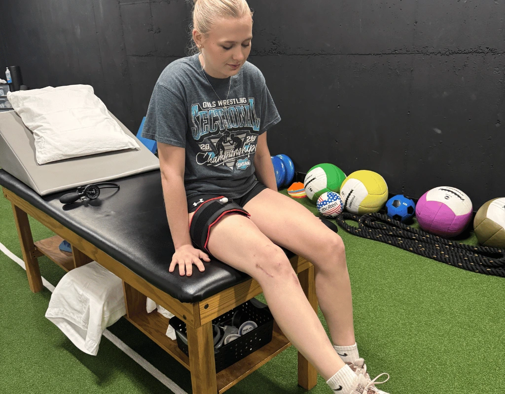 Patient seated on the edge of a treatment table with a blood flow restriction cuff strapped around her upper thigh, medicine balls behind her