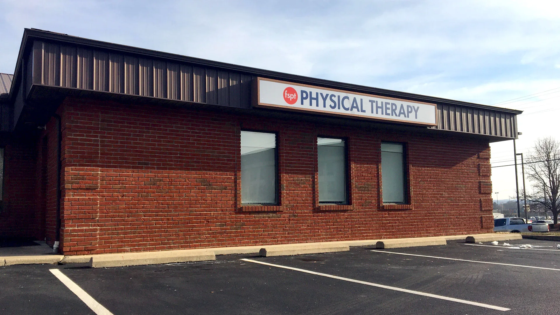Red brick building with a metal awning and a Physical Therapy sign above the windows, parking lot in the foreground