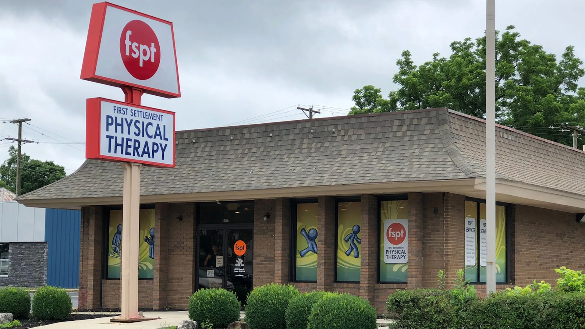 Brick clinic with a shingled roof and a tall red pole sign carrying the clinic logo and Physical Therapy lettering