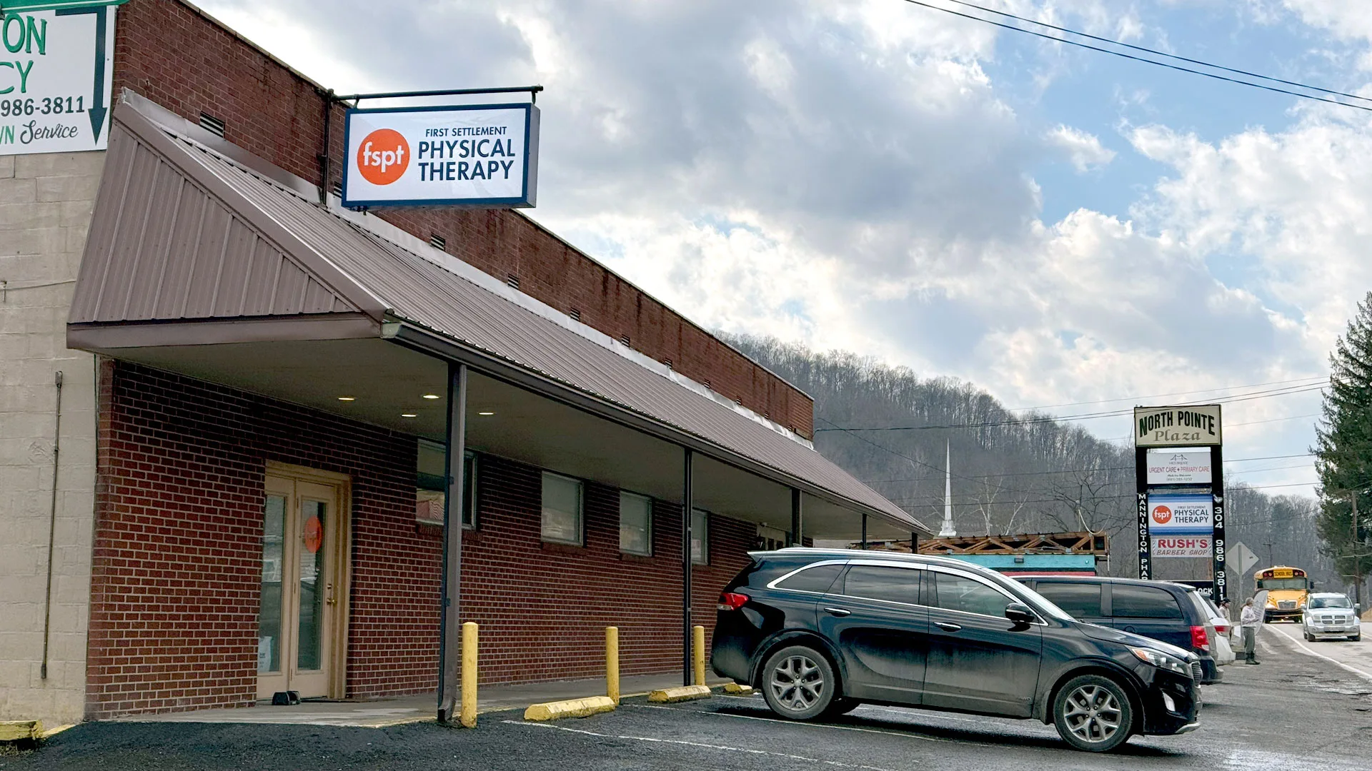 Brick building with a First Settlement Physical Therapy sign on the facade, cars parked alongside and a hillside behind