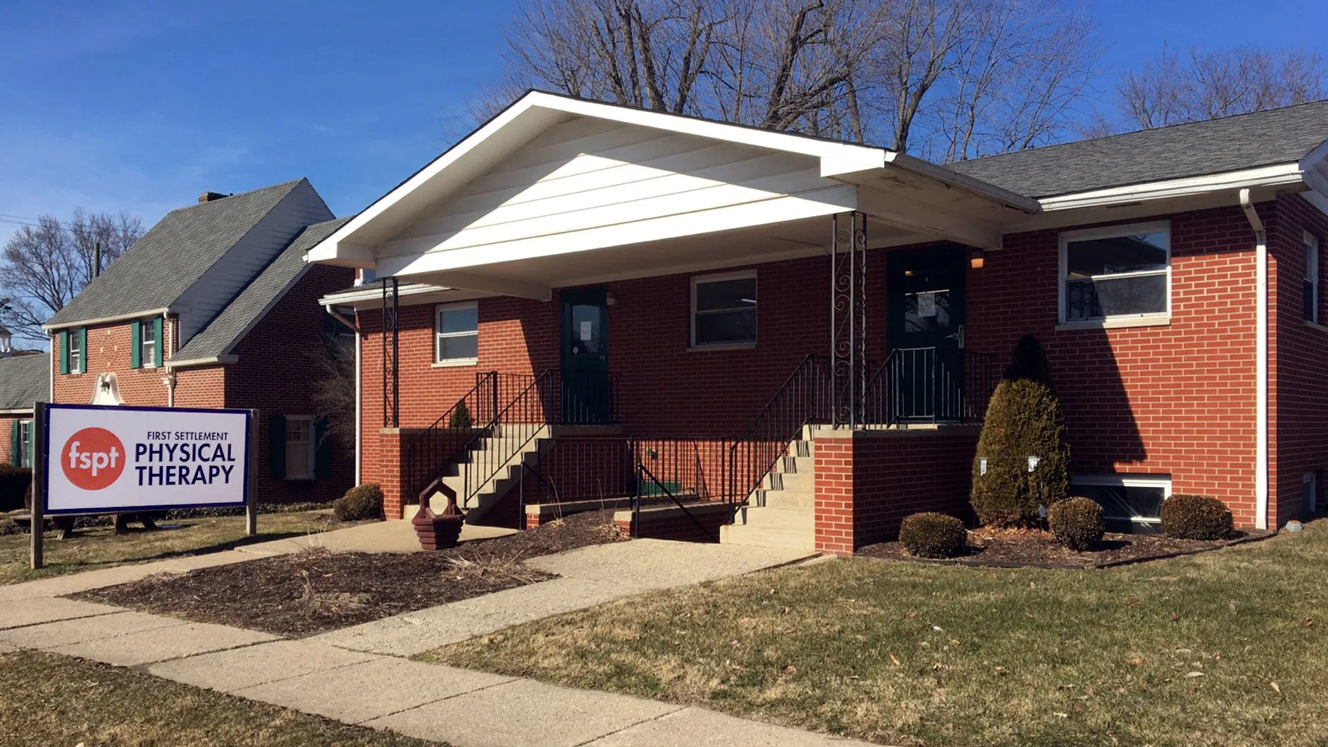 Brick house-style clinic with a small covered porch, entrance steps and a First Settlement Physical Therapy sign in the yard