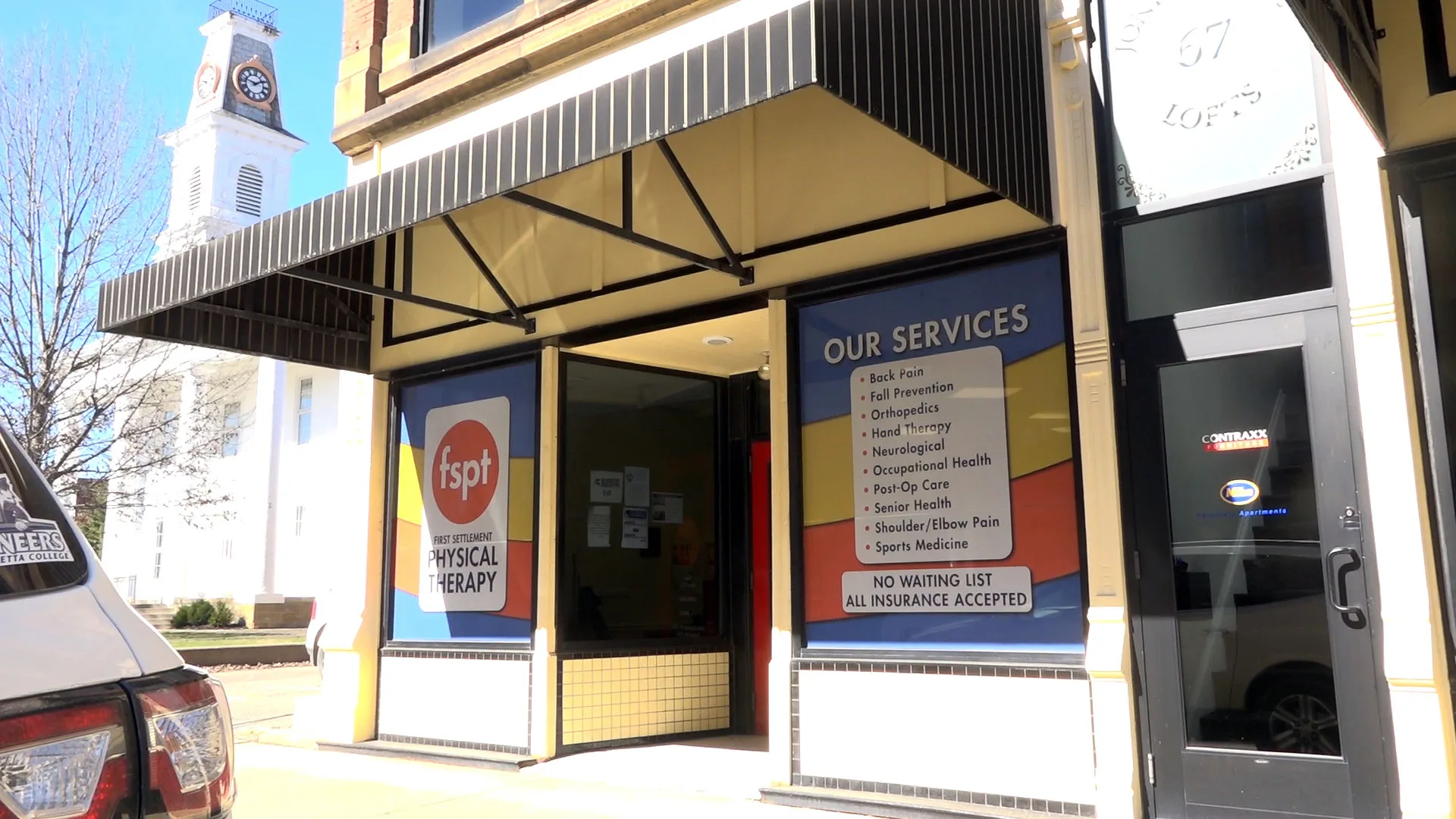 Storefront beneath an awning with an Our Services window sign, a clock tower visible on the building behind