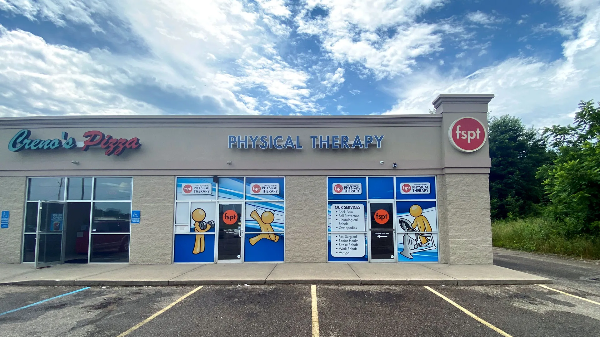Plaza storefront with the physical therapy unit and clinic logo sign alongside neighbouring businesses and a parking lot