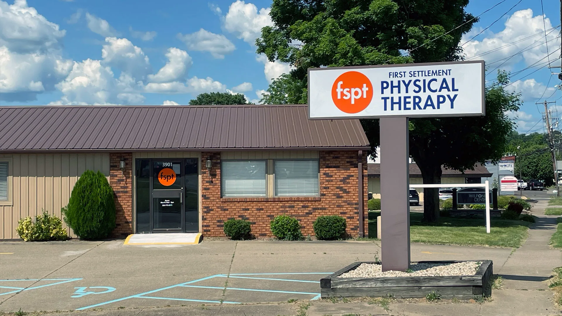 Small brick clinic building with a tall pole sign reading First Settlement Physical Therapy and trimmed shrubs along the walk