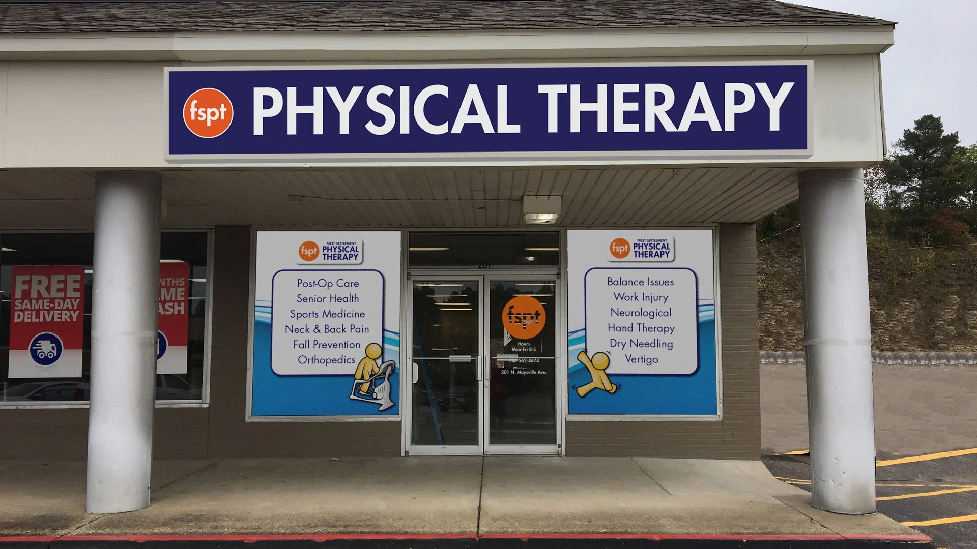 Clinic entrance framed by white columns with a blue Physical Therapy sign above the doors and service decals on the glass