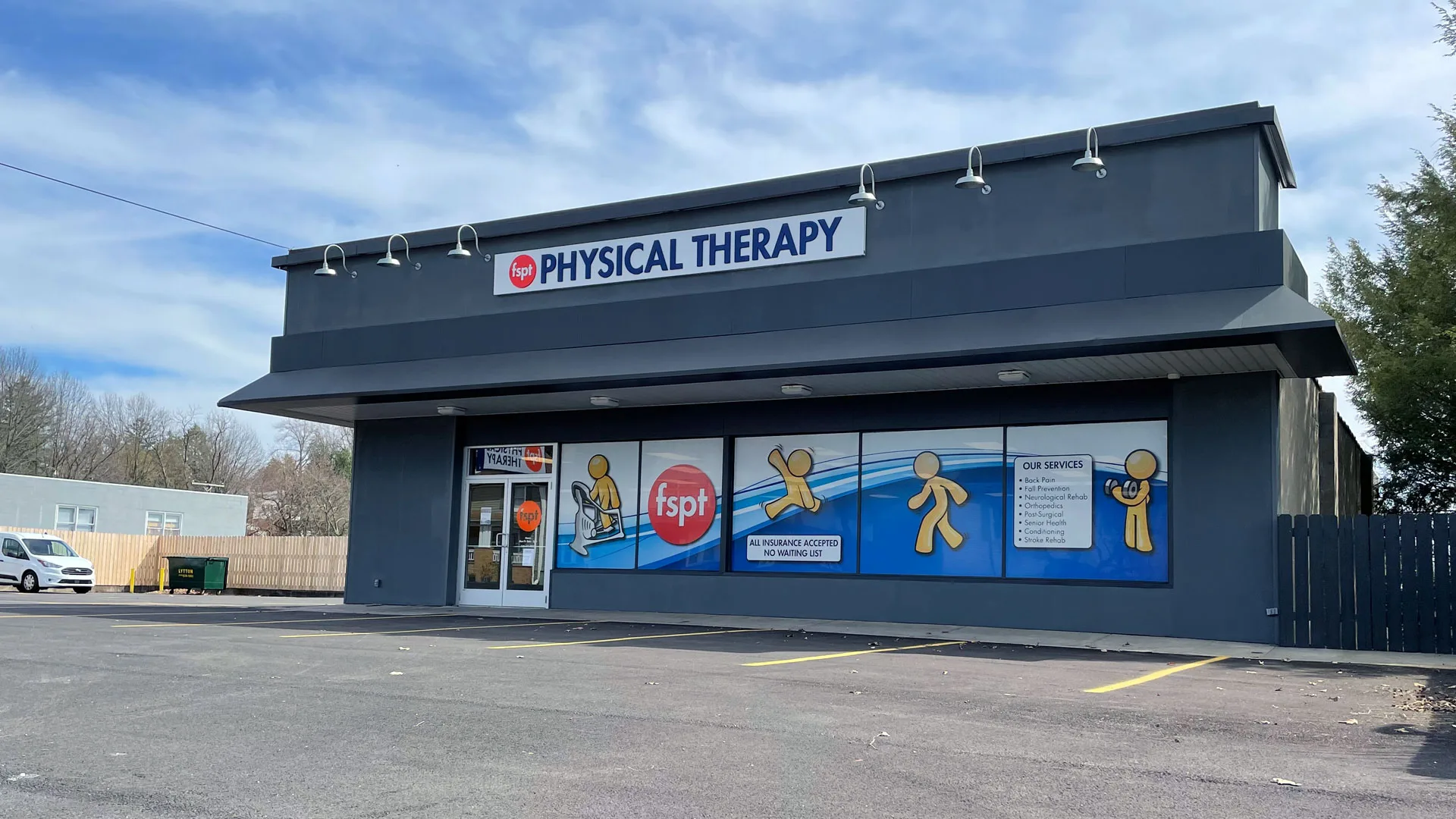 Standalone clinic with a dark roof, a blue Physical Therapy sign band and window decals, fronted by a large parking lot