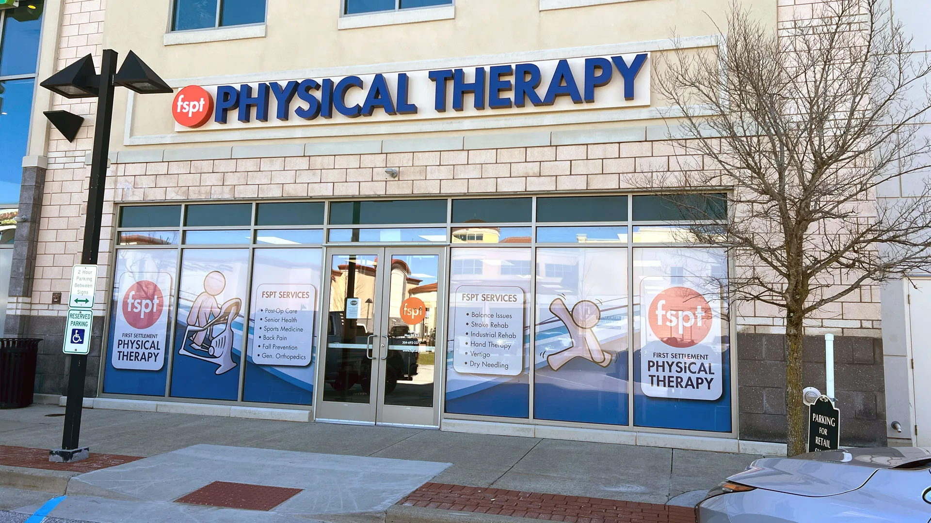 Corner clinic with a large First Settlement Physical Therapy sign, glass storefront covered in service decals and a bare tree outside