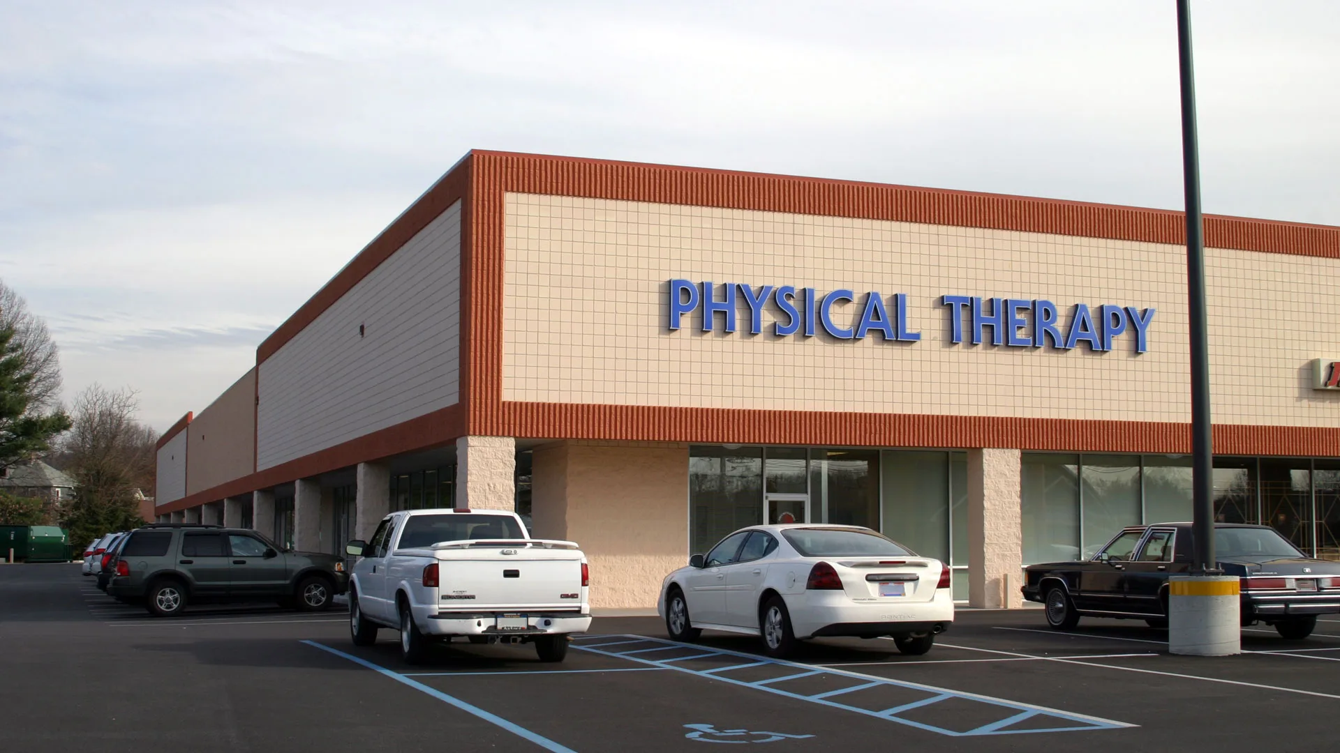 Large tan shopping-center building with blue Physical Therapy lettering above the storefront and cars parked in front