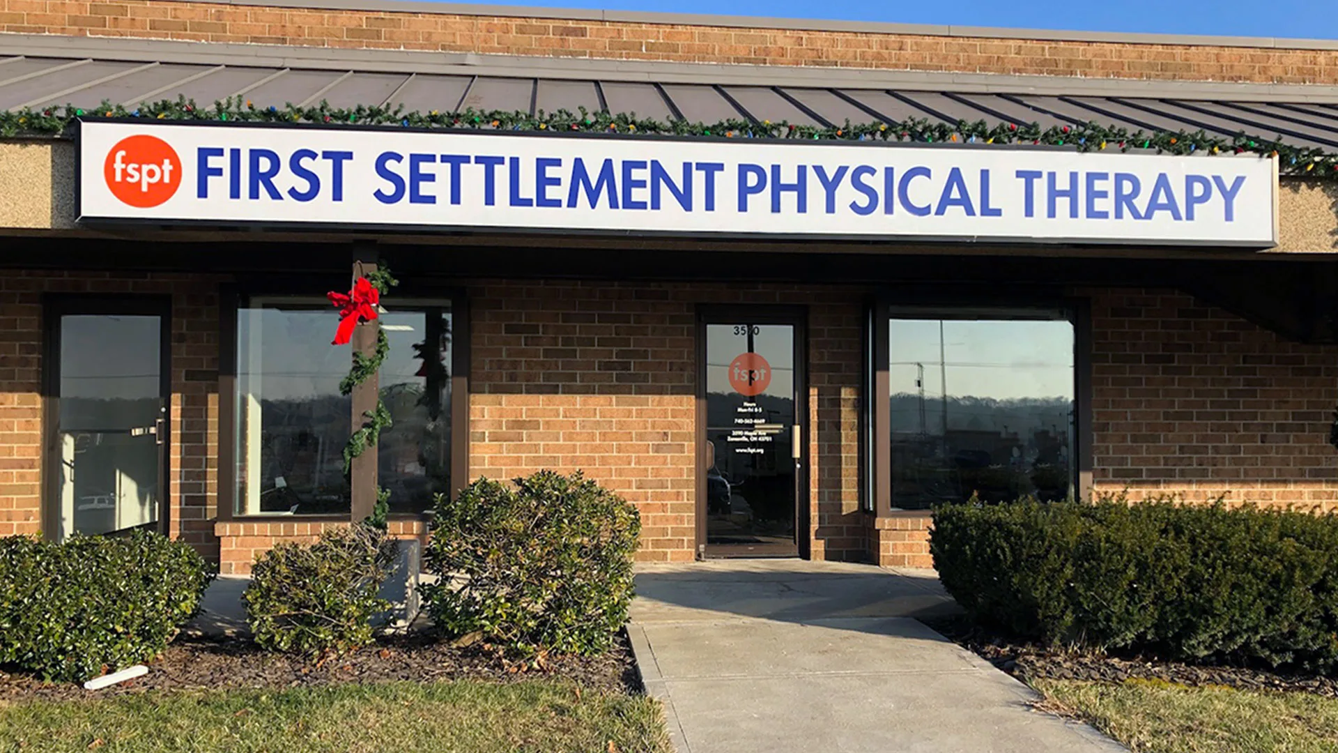 Brick clinic exterior with a large First Settlement Physical Therapy sign above the windows and a holiday bow on a lamppost